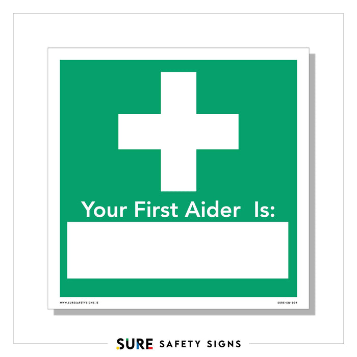 A green and white safety sign features a prominent medical cross above the customizable text Your First Aider Is: leaving a blank space for adding a name.