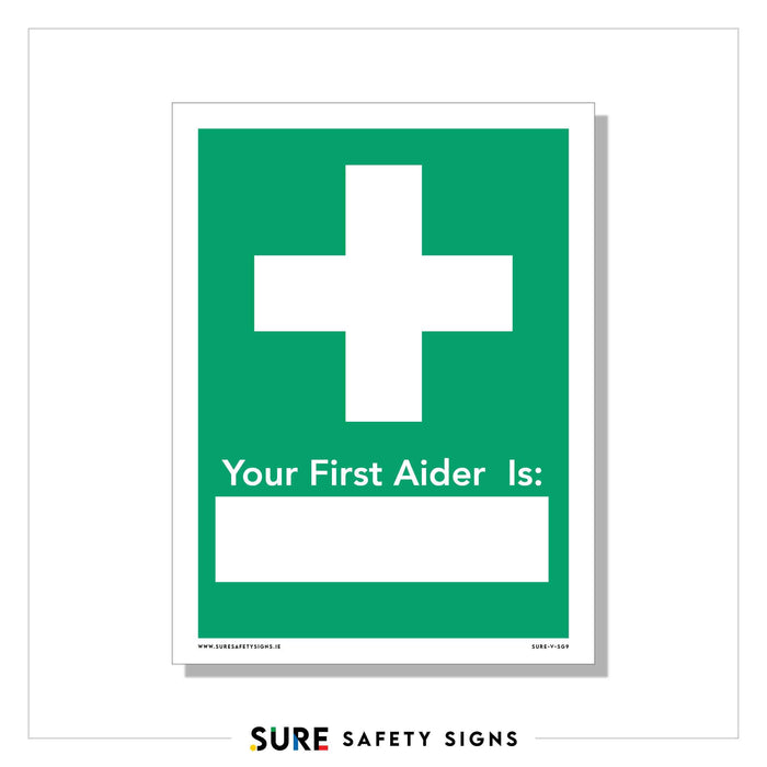 A green and white customizable safety sign features a prominent white cross, indicating emergency first aid information. Below, it reads Your First Aider Is: with space to fill in a name, blending utility with clear safety communication.
