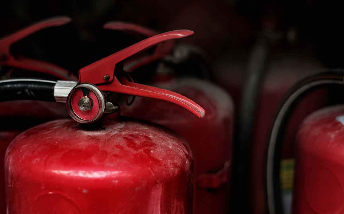 Close-up of a red fire extinguisher with a focus on the nozzle, pressure gauge, and handle. Other fire extinguishers are blurred in the background, creating a sense of depth. The extinguisher appears slightly worn, indicating regular use or handling.