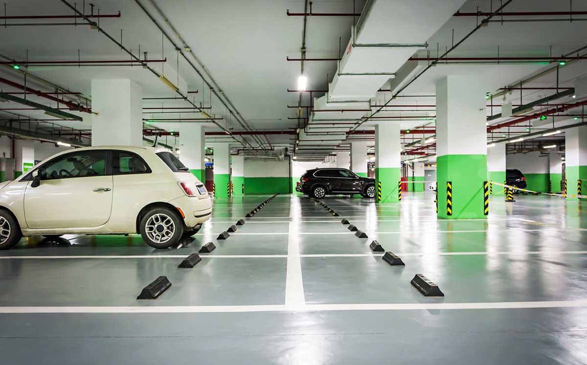 An underground parking garage with bright lighting. A white compact car and a black SUV are parked in marked spaces. Green and white columns are seen, and the floor and ceiling have visible pipes and signs.