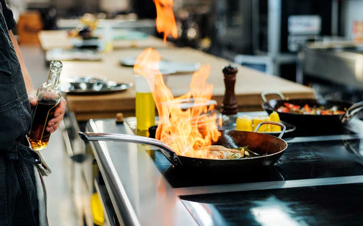 Chef holding a bottle of oil while a pan on the stove is aflame, suggesting flambé cooking. Surrounding the scene are ingredients and uncooked corn on the cob, with other pans in the background.